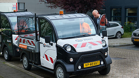 Man in pak bekijkt een compact busje met laadbak, zwaailicht en rode strepen. Op het nummerbord staat "vpwerkt".