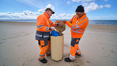 Twee mensen in oranje werkkleding legen een prullenbak met het strand op de achtergrond.