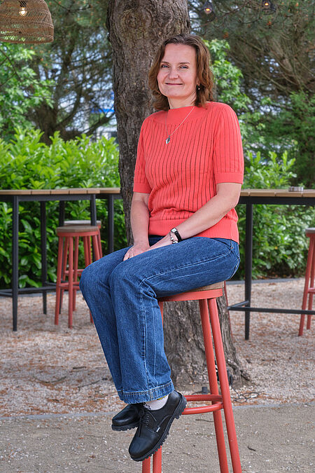een vrouw poseert op een kruk voor een boom en veel groen op de achtergrond.