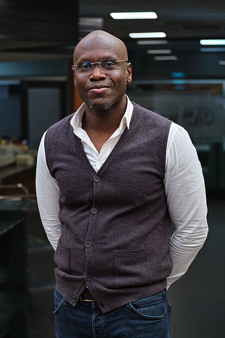 poserende man in een net grijs hesje, wit shirt en een spijkerbroek.