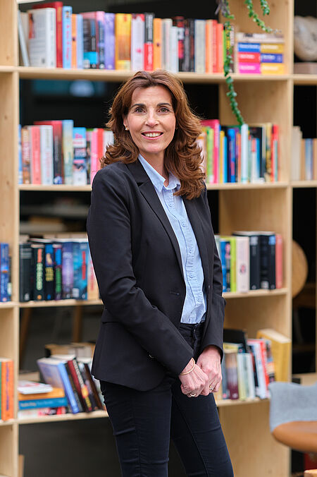 vrouw met lang bruin haar en nette kleding poseert voor een boekenkast