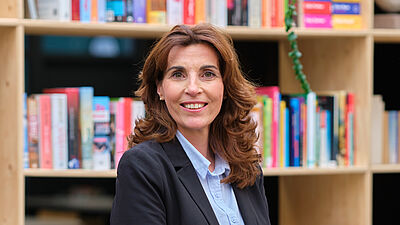 vrouw met lang bruin haar en nette kleding poseert voor een boekenkast