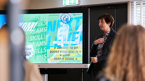 Vrouw spreekt mensen toe naast een presentatiescherm.