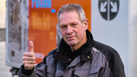 Portret van een man in werkkleding die zijn duim opsteekt. Vrachtwagen van VPW op de achtergrond.