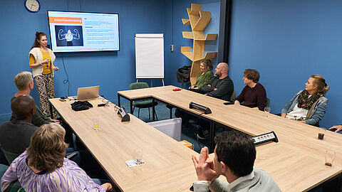 Vrouw geeft een presentatie in lesruimte met verschillende mensen rondom een tafel.
