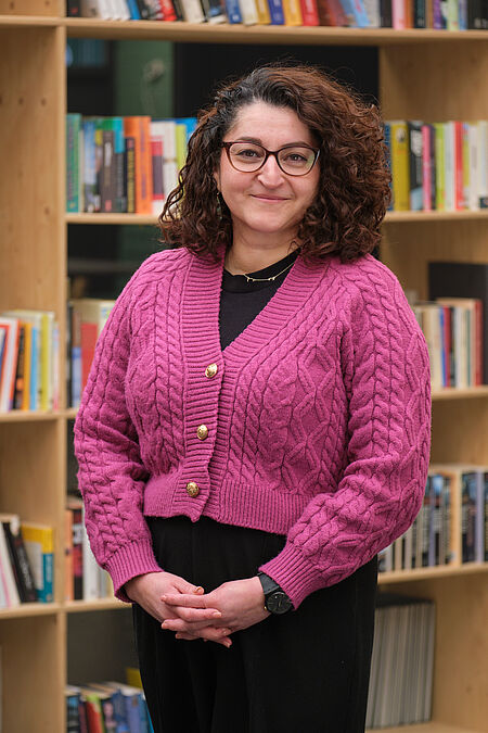 vrouw met krullen, een bril en een roze trui poseert voor een boekenkast