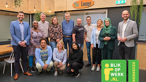 Groep van 13 diverse mensen in een ruimte met bordje van "The Lounge". Logo Blik op Werk rechtsonder in beeld.